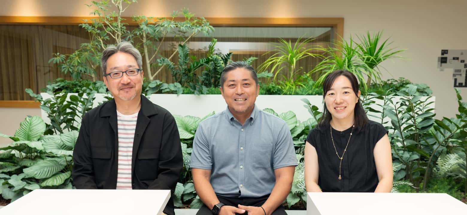 Group photo of three smiling employees sitting against a green background in the room.